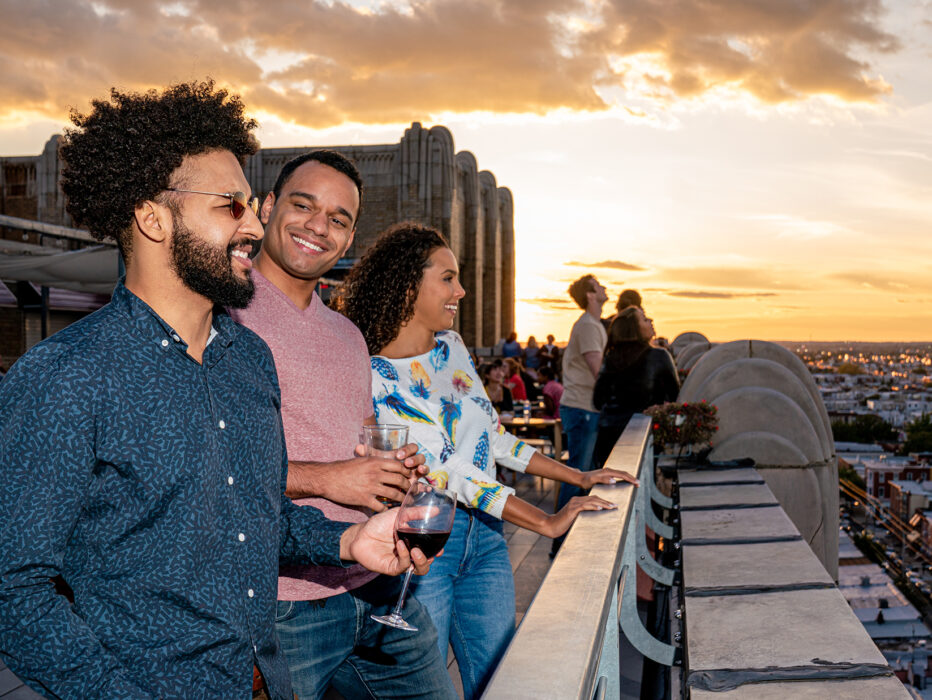 Three people enjoy drinks and look out at the view of the city while at Bok Bar rooftop during sunset. The sky illuminates in yellow and orange tones.