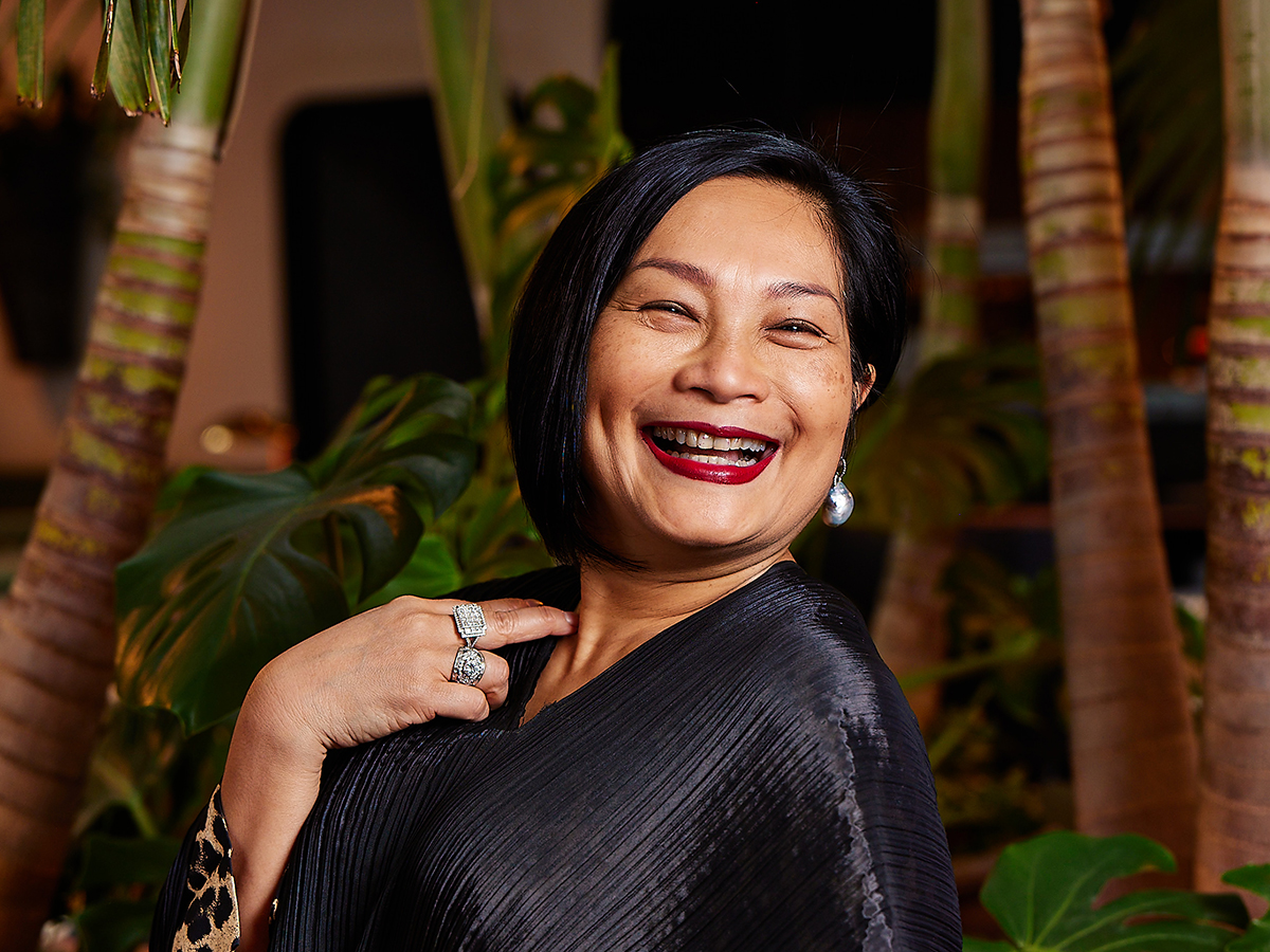 Le chef Nok Suntaranon sourit et pose pour une photo devant un décor tropical de palmiers.