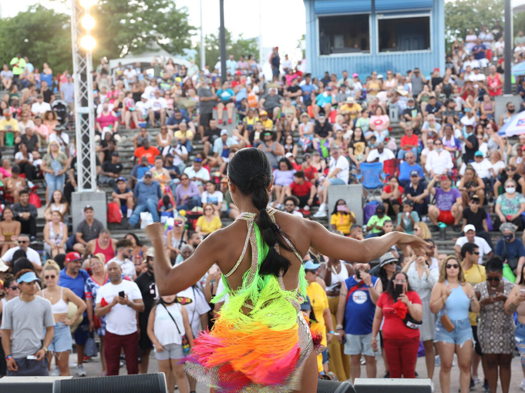 Une artiste vêtue d'une tenue à franges multicolores et lumineuses danse sur scène devant une foule nombreuse et enthousiaste.