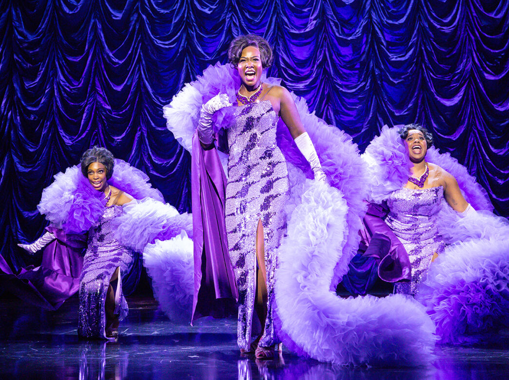 Three actresses wearing extravagant purple dresses with large tool capes sing and dance on stage during a performance of Dreamgirls.