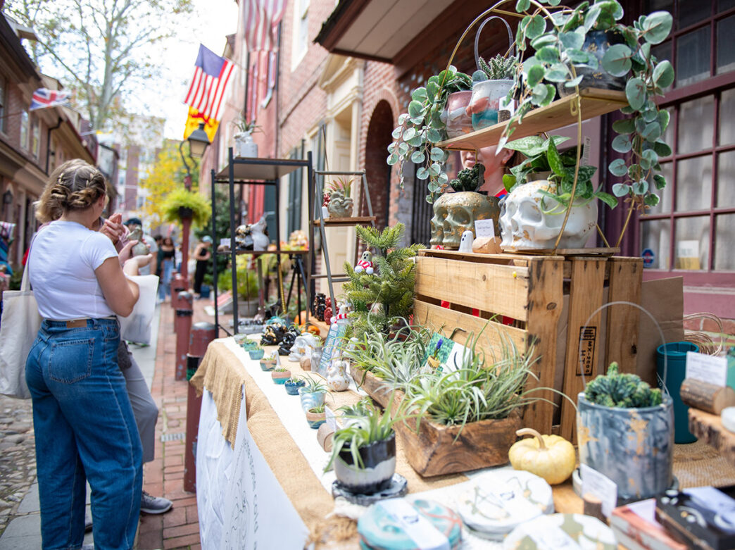 Crafts and goods are displayed on tables lining Elfreth's Alley for the Makers Market while people walk around and shop.