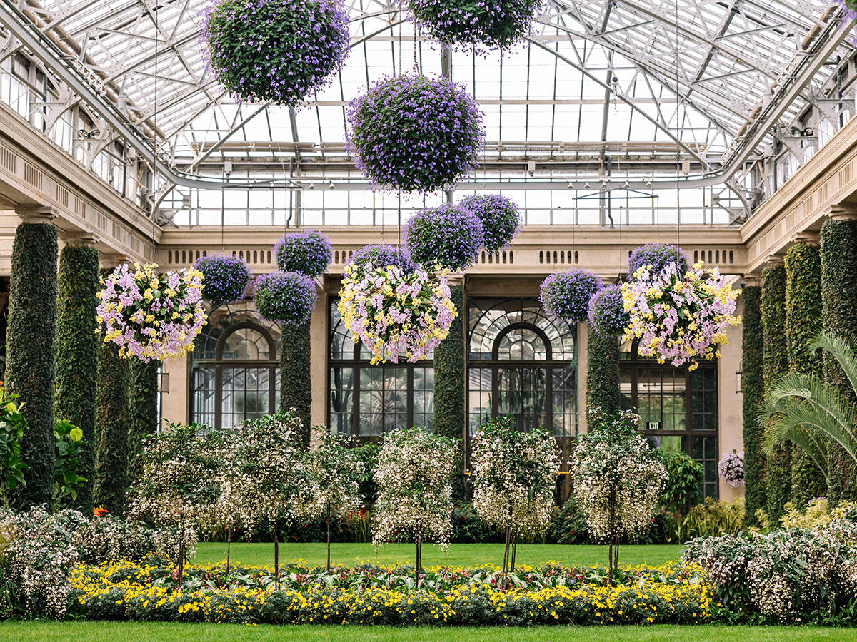 Um grande conservatório interno apresenta um telhado de vidro e colunas envoltas em hera. Grandes arranjos florais estão pendurados no teto.