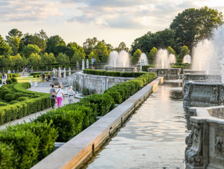 People walk through the Main Fountain Garden at Longwood Gardens. Green shrubs line the pathways between the spraying fountains.