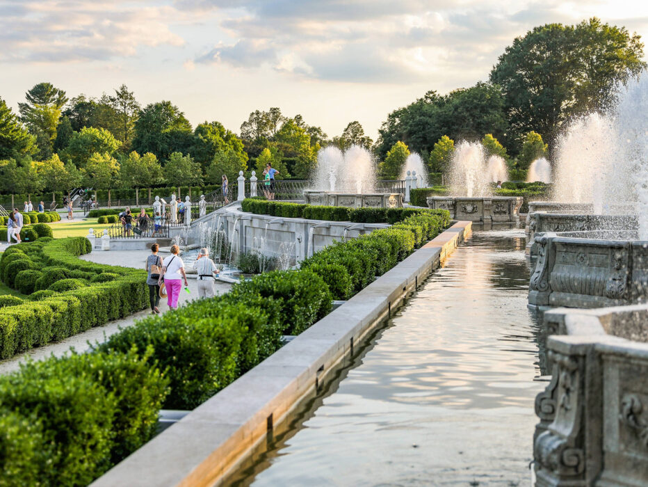 People walk through the Main Fountain Garden at Longwood Gardens. Green shrubs line the pathways between the spraying fountains.