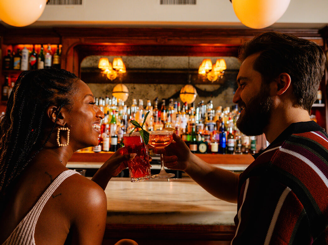Dos personas sentadas en una barra se enfrentan y brindan con sus cócteles.