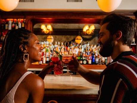 Two people seated at a bar face one another and toast their cocktails.