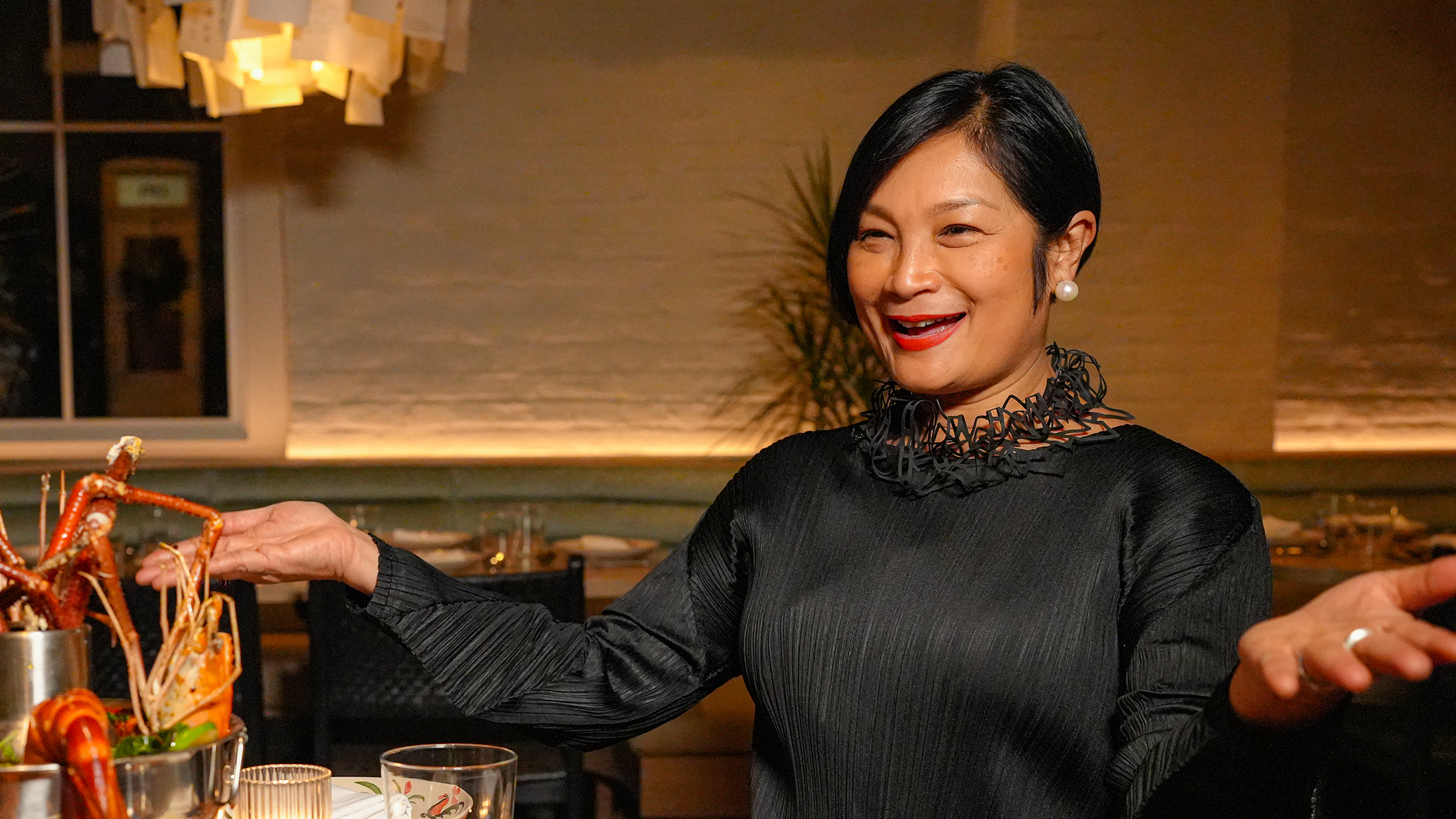 La chef Nok Suntaranon est assise à une table ronde en bois et tient les bras ouverts tout en présentant une tour de fruits de mer au Kalaya.