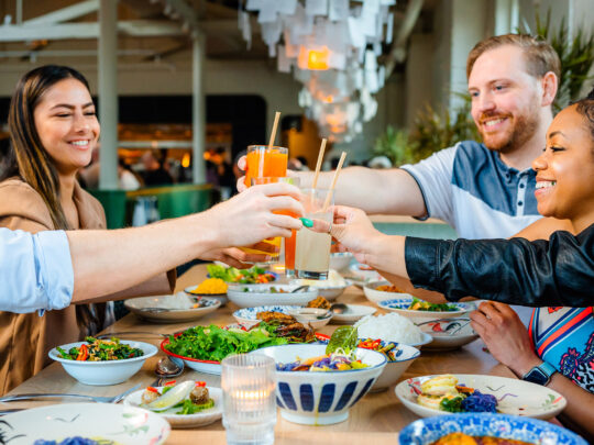 Un groupe d'amis souriants lèvent leur verre pour trinquer autour d'une longue table garnie de plats thaïlandais colorés.