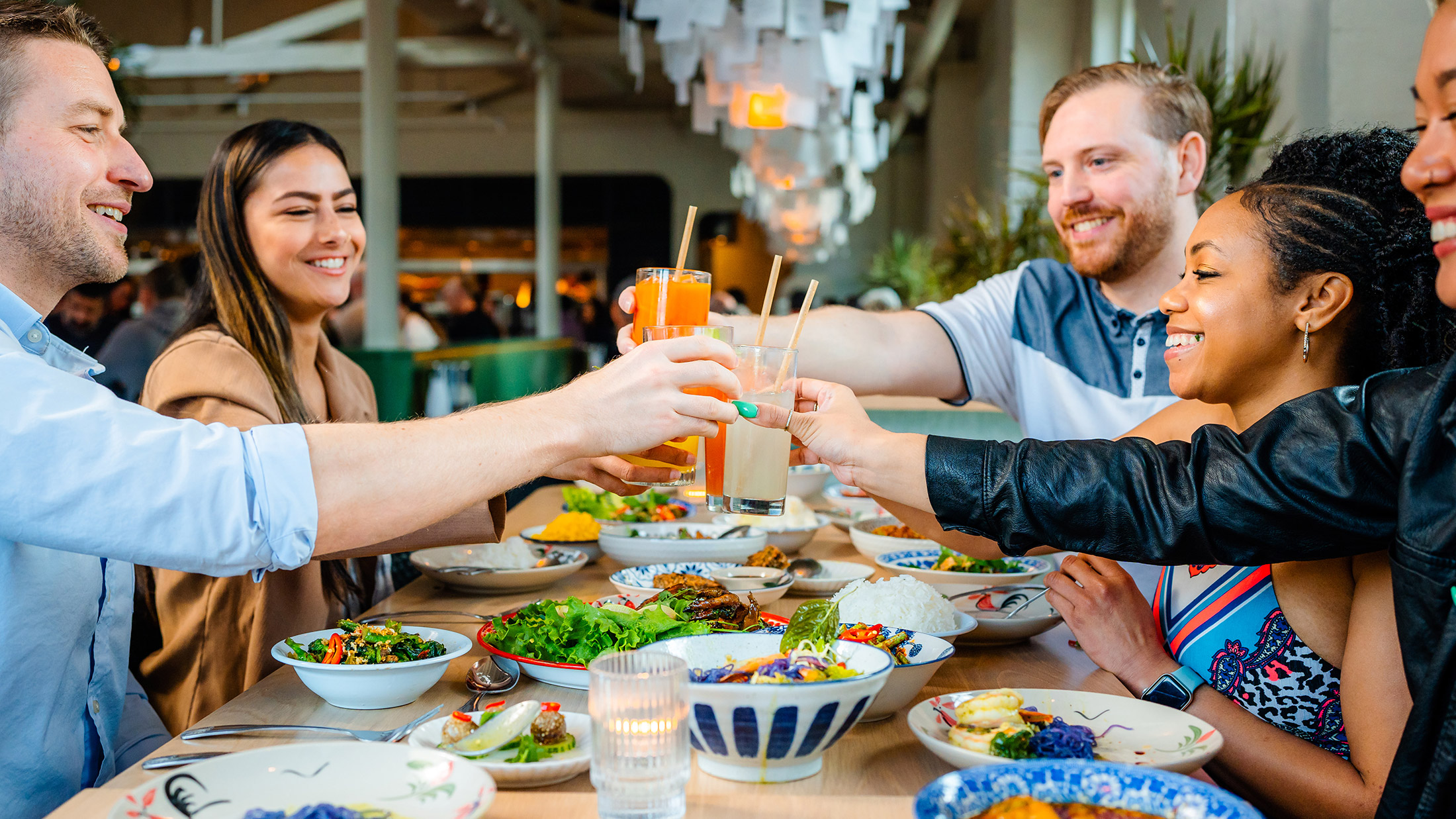 A group of smiling friends raise their glasses for a toast over a long table filled with colorful Thai cuisine.