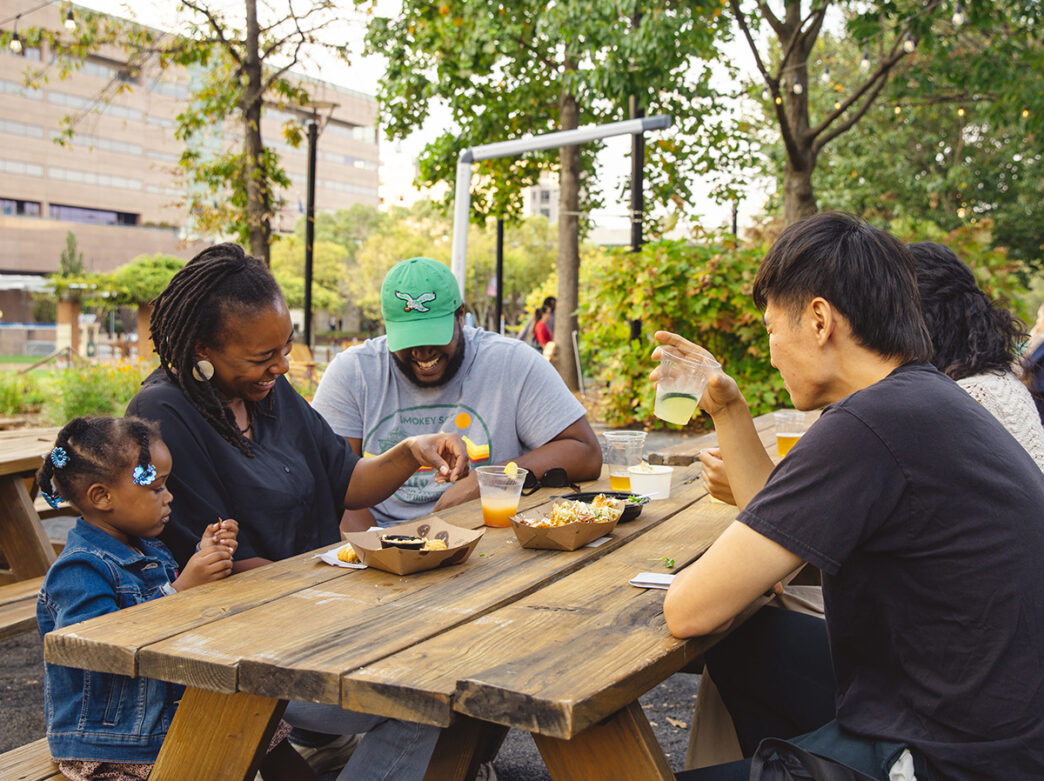 Four adults and one child sit at a picnic table and laugh while enjoying drinks and food items at Liberty Garden.