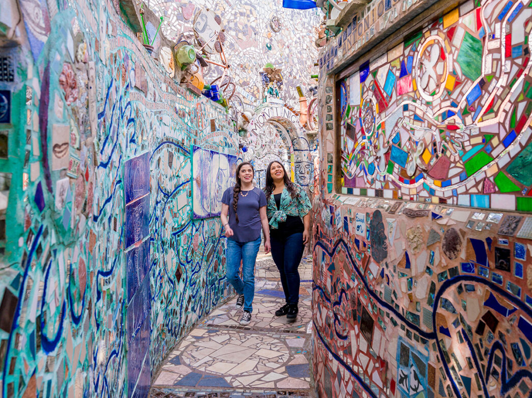 Two visitors walk through a narrow, winding walkway at Philadelphia’s Magic Gardens, surrounded by intricate mosaic walls adorned with colorful tiles, mirrors, bottles and found objects forming dazzling, abstract patterns.