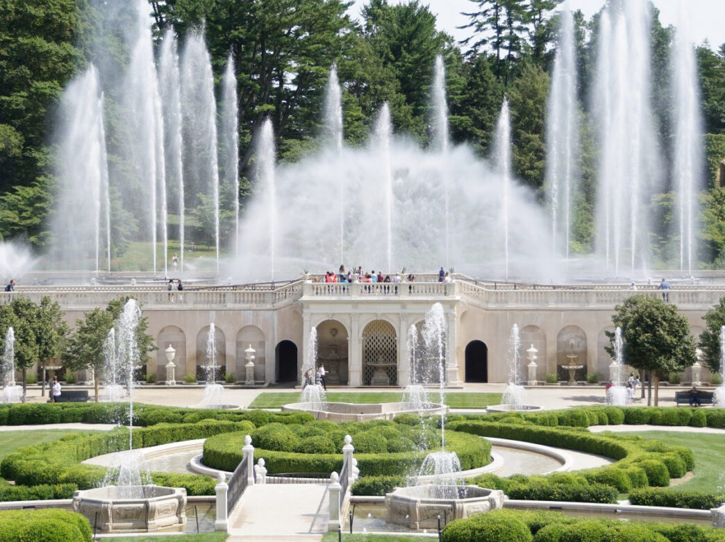A large, elegant courtyard with symmetrical fountains and trimmed hedges. Power water jets shoot water high into the air while people walk through the garden space.