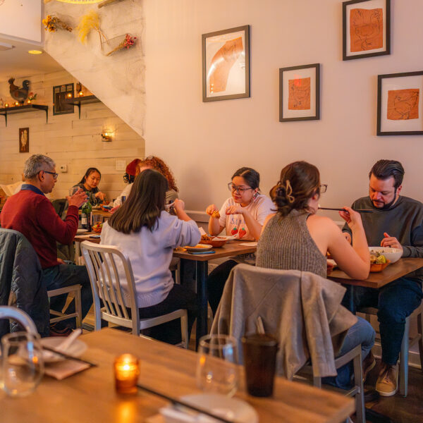 A warm, inviting dining room at Mawn with guests seated at small wooden tables, enjoying their meals. Drawings of chickens on brown paper are displayed in frames on the walls.
