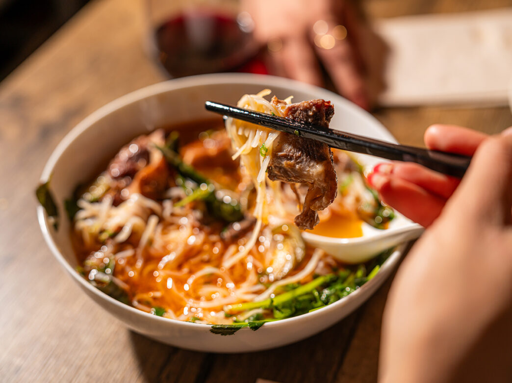 A close-up shot of a bowl of Cambodian noodle soup, featuring thin rice noodles, slices of beef and herbs. A hand holding chopsticks lifts a piece of the beef and noodles from the bowl.