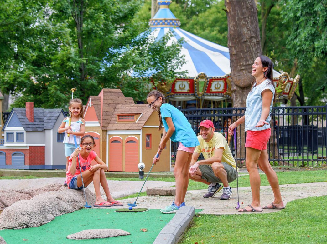 A smiling family plays a round of mini golf surrounded by miniature houses, with a colorful carousel peeking through the trees in the background.