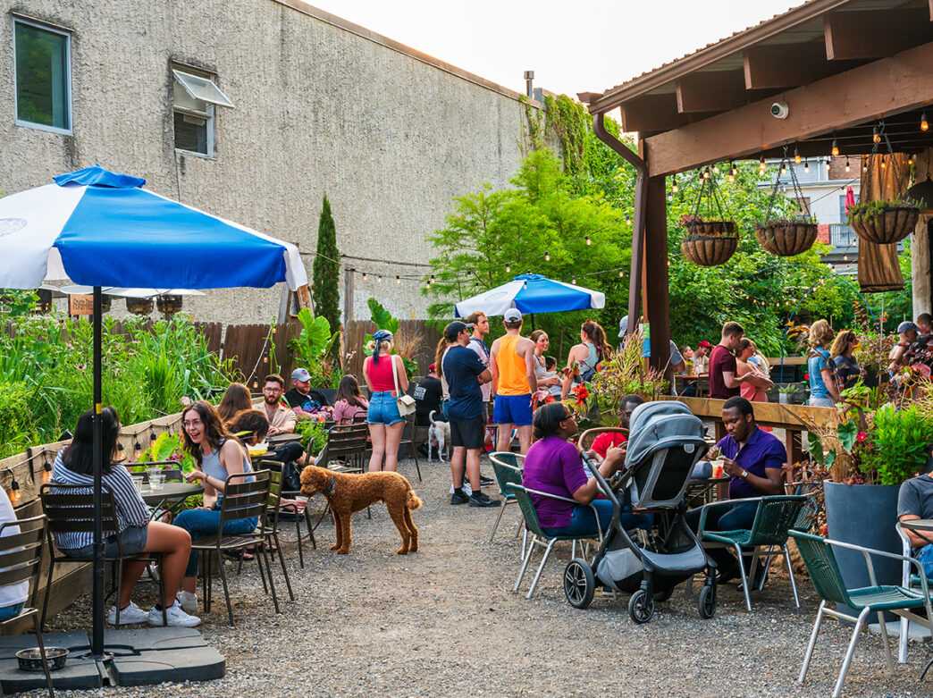 People enjoy drinks and bites in a lush garden setting at the PHS Pop Up Garden on South Street. The garden is complete with shaded tables and hanging planters.
