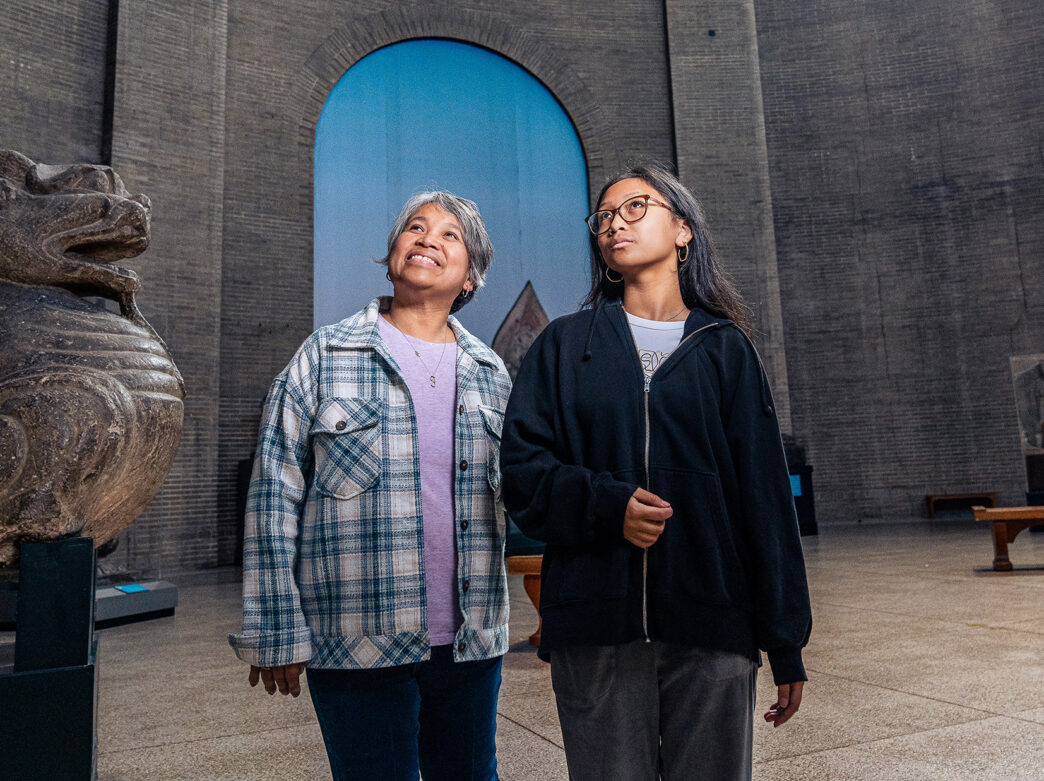 Two visitors walk through the rotunda at the Penn Museum, passing an ancient stone sculpture, with the museum's high-arched walls and intricate windows in the background.