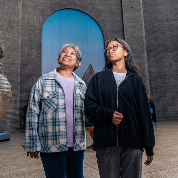 Two visitors walk through the rotunda at the Penn Museum, passing an ancient stone sculpture, with the museum's high-arched walls and intricate windows in the background.