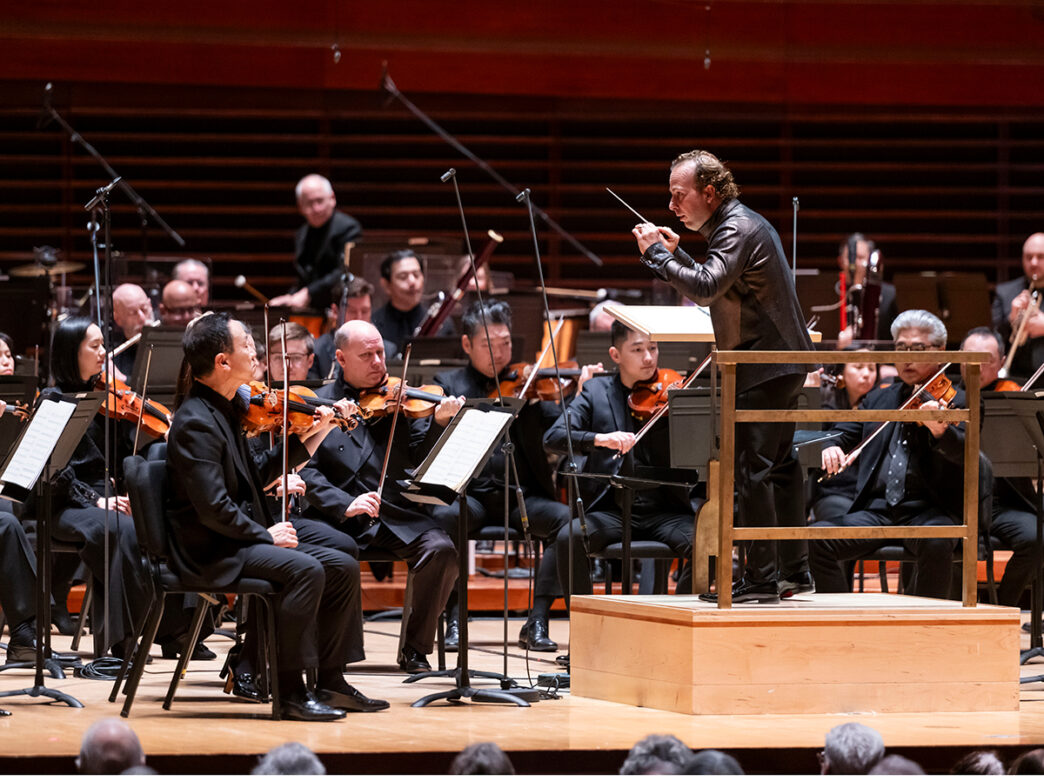 Composer Yannick Nézet-Séguin stands on a platform in front of the string section of the orchestra.