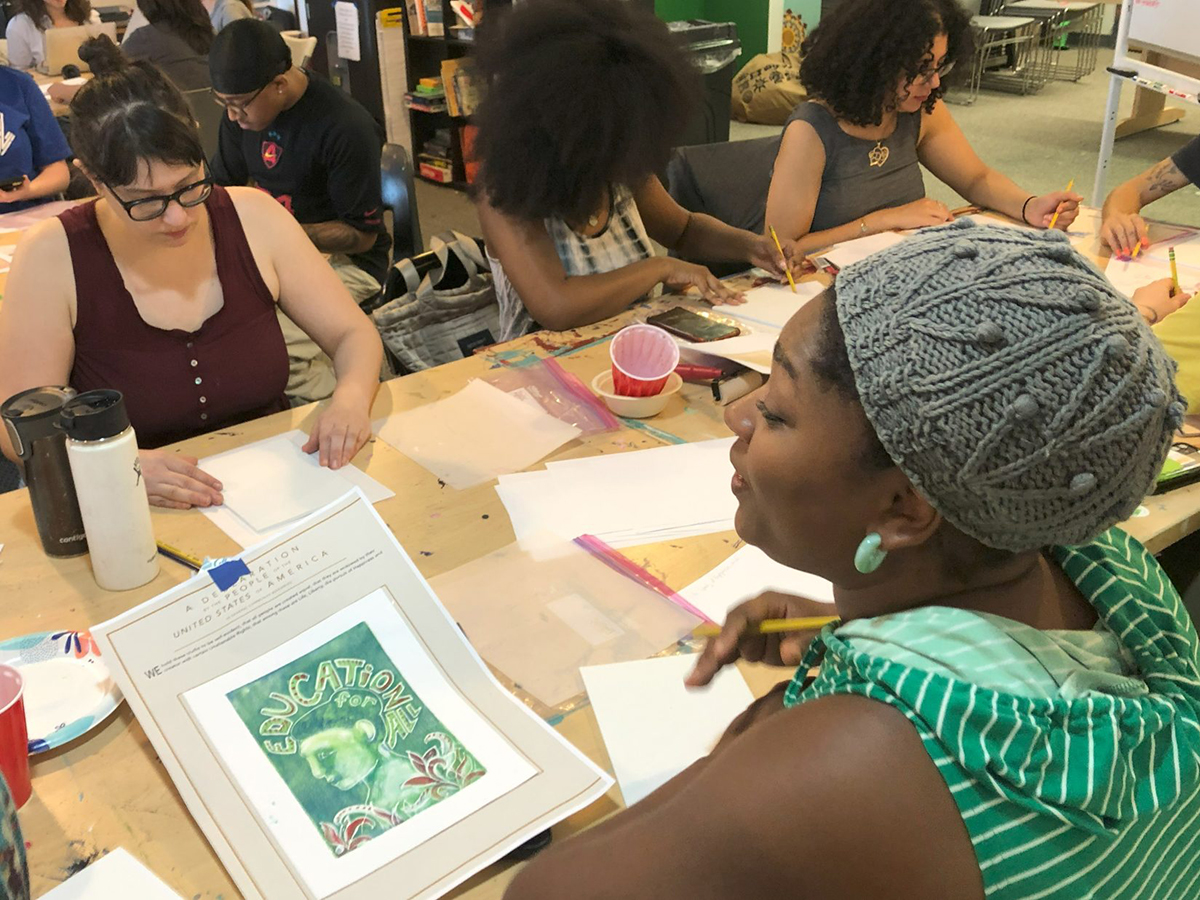 Participants working on art projects during a printmaking workshop are gathered around a table with paper, pencils and artwork.
