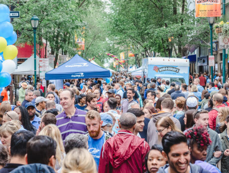 Crowds of people walk down Walnut Street. Someone on the left carries and arrangement of blue, white and yellow balloons, and vendor tents and food trucks are set up in the street.