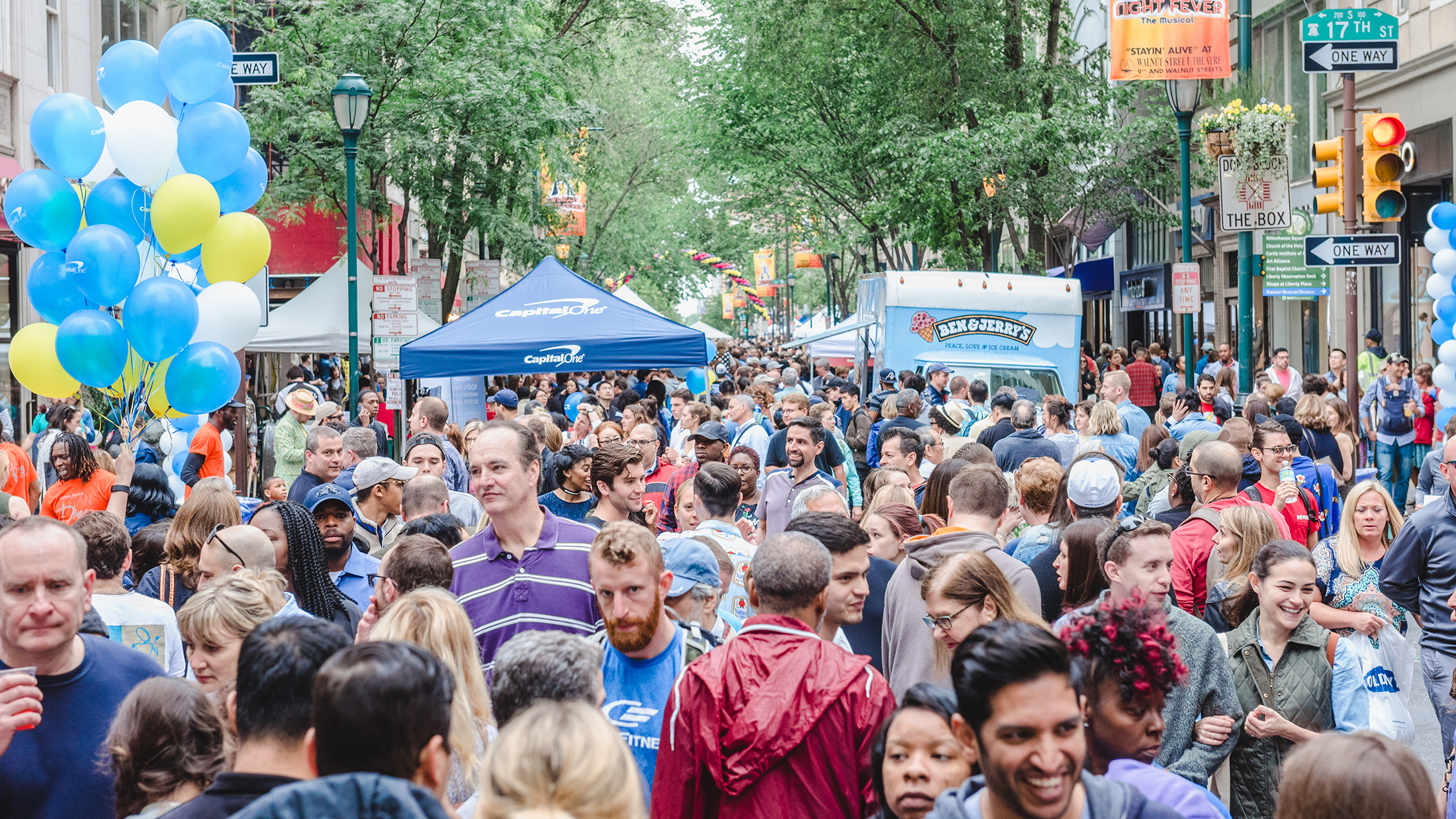 Crowds of people walk down Walnut Street. Someone on the left carries and arrangement of blue, white and yellow balloons, and vendor tents and food trucks are set up in the street.