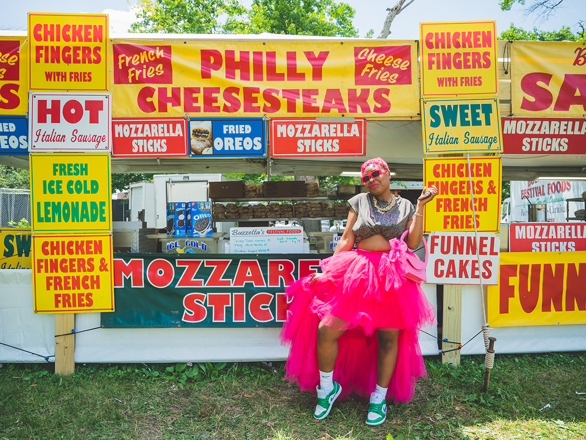 A person wearing a hot pink tool skirt, pink sunglasses and green sneakers poses for a phot in front of a food stand with cheesesteaks, chicken fingers, mozzarella sticks and more.