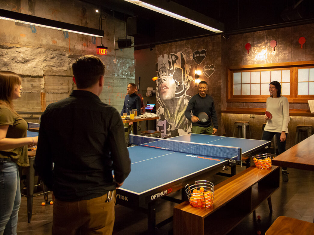 A group of friends plays ping-pong at SPIN Philadelphia. The group laughs and enjoys the vibrant, industrial-chic space decorated with colorful murals and playful paddle-themed art.