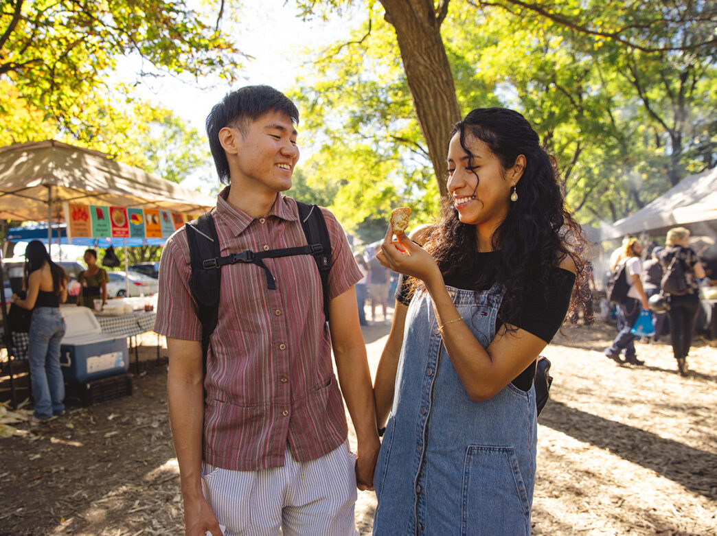 Un couple souriant se tient la main en se promenant dans le marché du Sud-Est asiatique dans le parc FDR. La femme, vêtue d'un pull en jean, brandit une boulette et sourit à son compagnon dans un décor animé de tentes et d'arbres.