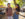 A smiling couple holds hands while strolling through the Southeast Asian Market in FDR Park. The woman, wearing a denim jumper, holds up a dumpling and smiles at her partner amid a lively backdrop of food tents and trees.