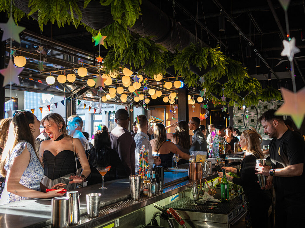 People stand at the bar, socialize and drink cocktails at Stratus Rooftop Lounge. The bar is decorated with hanging greenery, silver stars and glowing orbs.