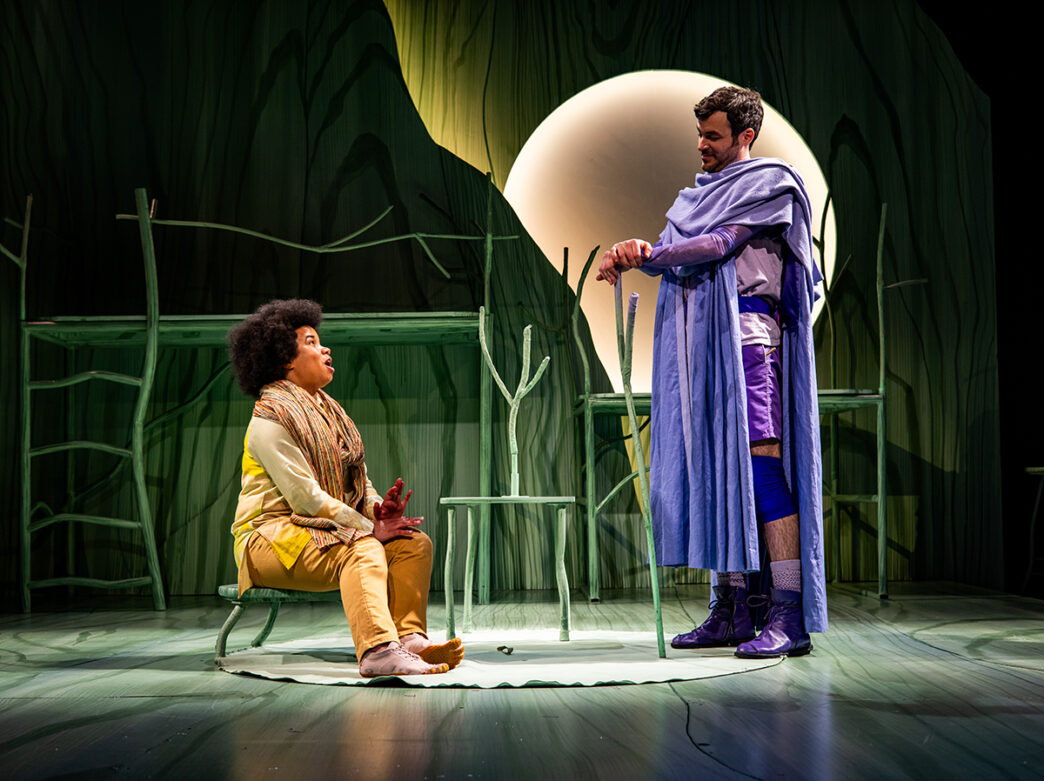 An actor portraying Bilbo sits on a small green stool, while another actor playing Gandalf stands beside him in a long purple cloak and boots. The stage glows under soft stage lighting, with a large illuminated full moon in the background.