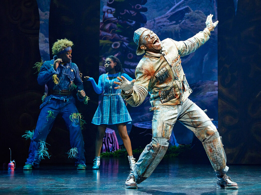 Trois acteurs se produisent sur scène dans une vibrante scène de The Wiz. L'un d'eux, déguisé en homme de fer blanc, se tient sous les projecteurs, les bras écartés et la bouche ouverte en chantant, tandis que les deux autres, déguisés en épouvantail et en Dorothy, réagissent de manière expressive derrière lui.