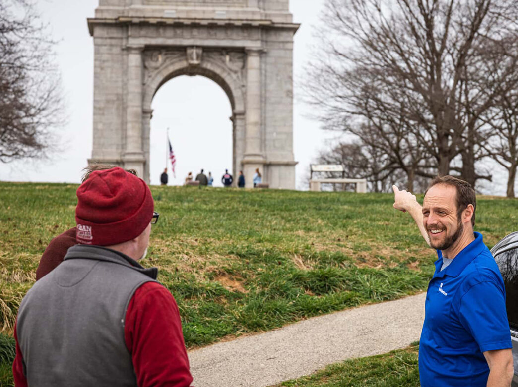 A guide wearing a blue polo points to a large marble arch at Valley Forge National Historical Park.
