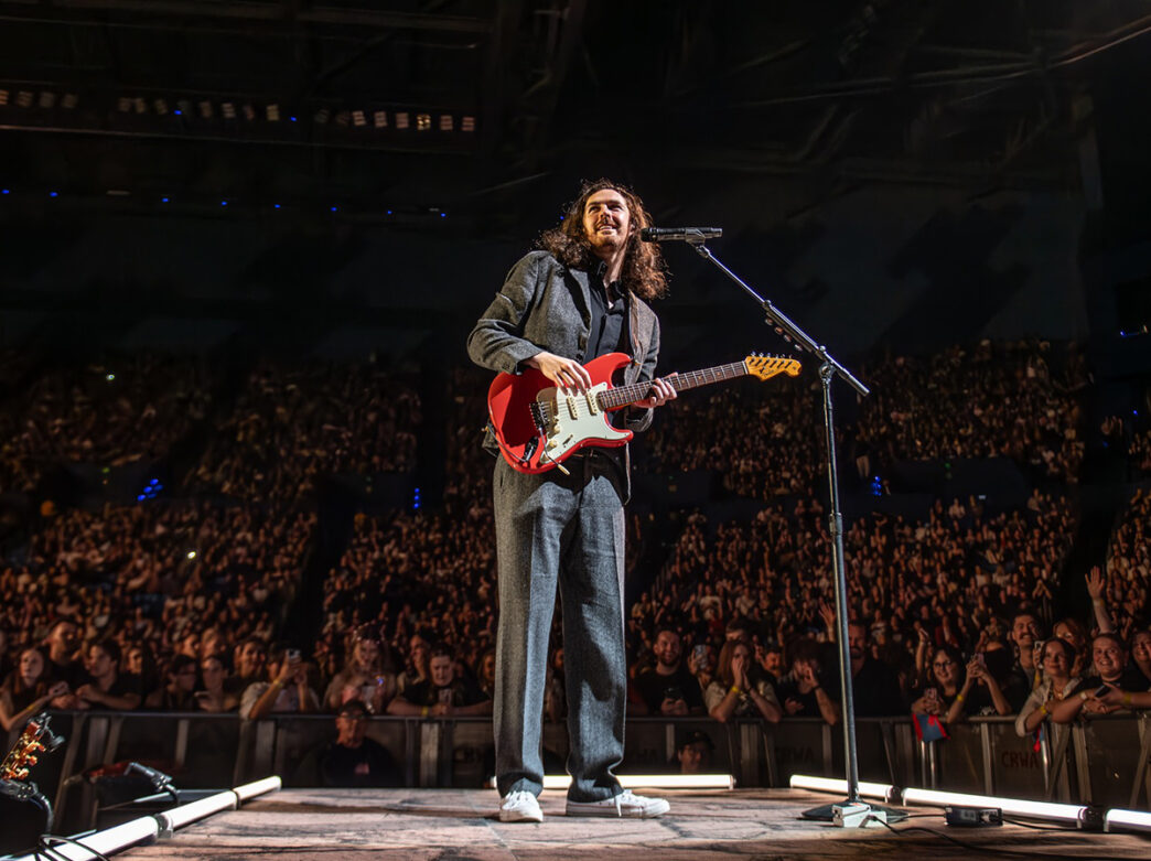 Le musicien Hozier porte un costume gris et des baskets blanches tout en jouant d'une guitare électrique rouge sur scène. Derrière lui, on voit une foule qui remplit un stade.