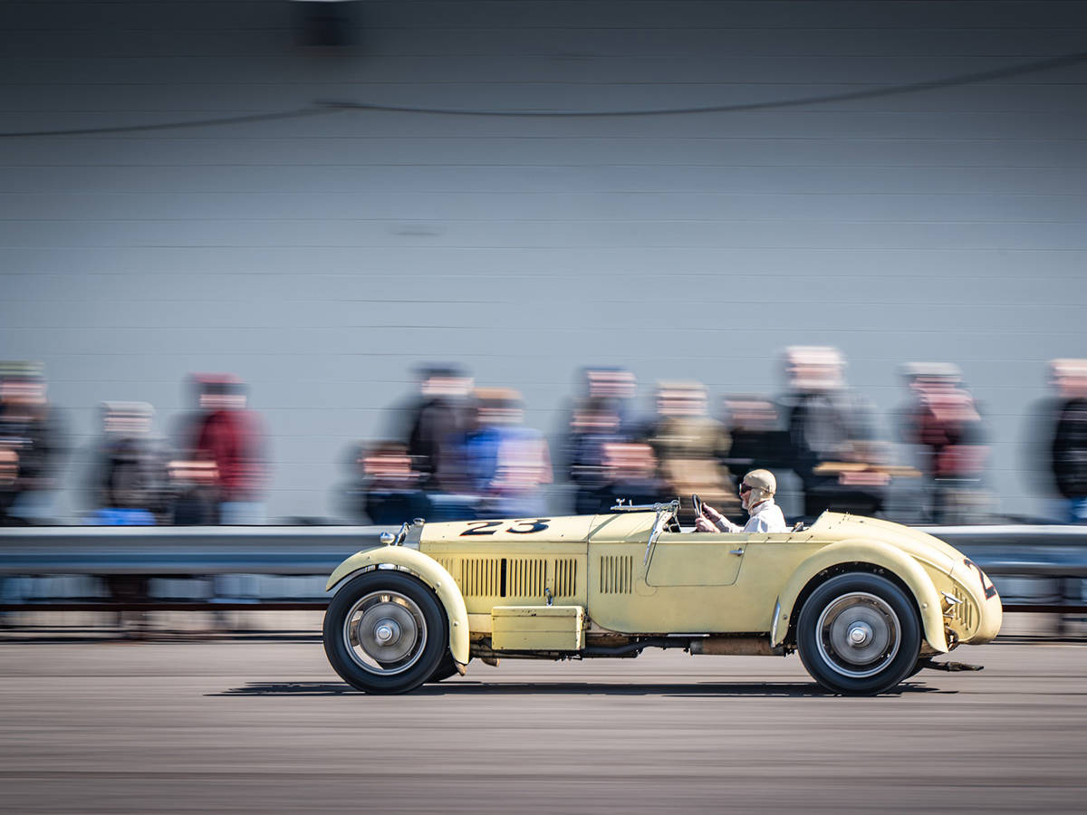 A yellow vintage racing car marked “23” speeds past a blurred crowd during a demonstration.