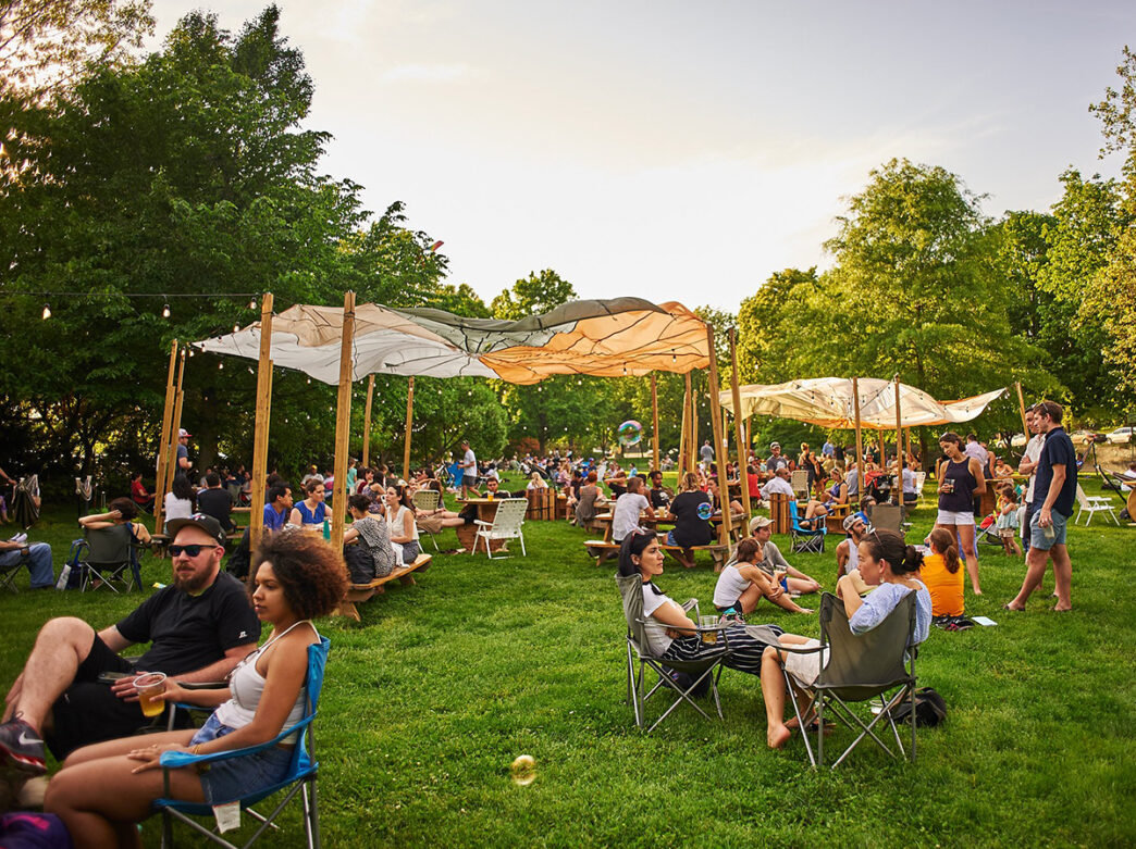 Une foule de personnes sont assises sur des chaises de camping et des tables de pique-nique, sous des parasols, sur une pelouse du parc Fernhill. Des bulles flottent dans l'air.