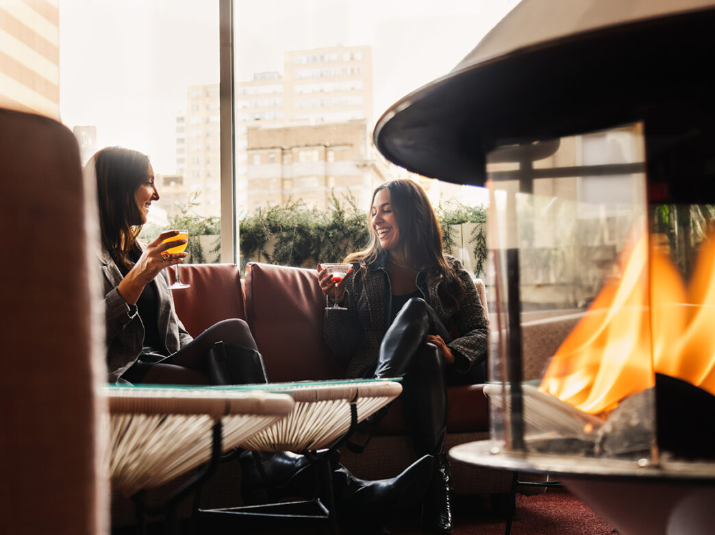 Two people sit on a couch while drinking cocktails at the Continental Midtown's rooftop lounge. A firepit glows with orange flames.
