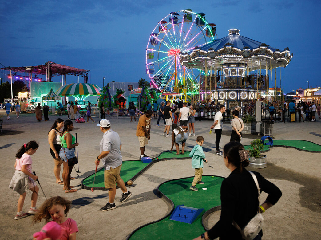 Adults and children play mini-golf in front of a Ferris wheel, carousel and other amusement rides at the Independence Blue Cross River Rink Summerfest.