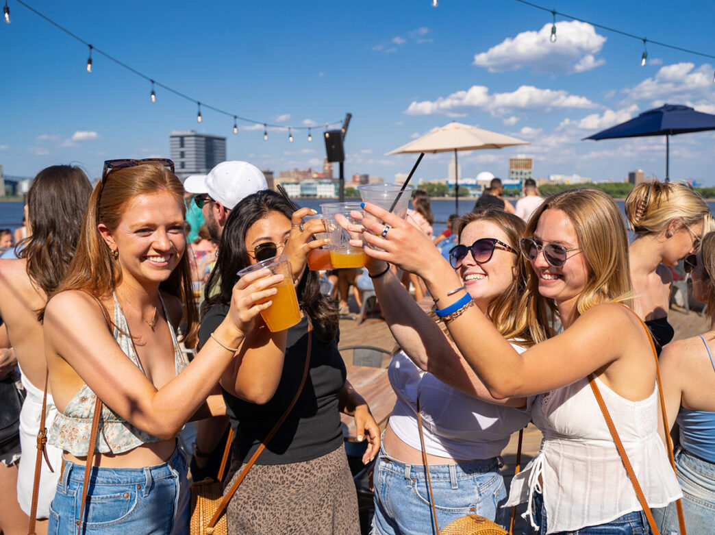 A group of friends raises their drinks for a cheers under sunny skies at Liberty Point, with the Delaware River and string lights in the background.