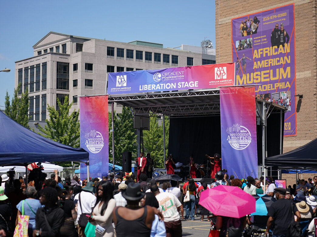 Une foule se rassemble à l'extérieur du African American Museum in Philadelphia pour une fête de quartier animée à l'occasion du dix juin. Les gens regardent des artistes en tenue rouge chanter sur une scène ornée de bannières colorées.