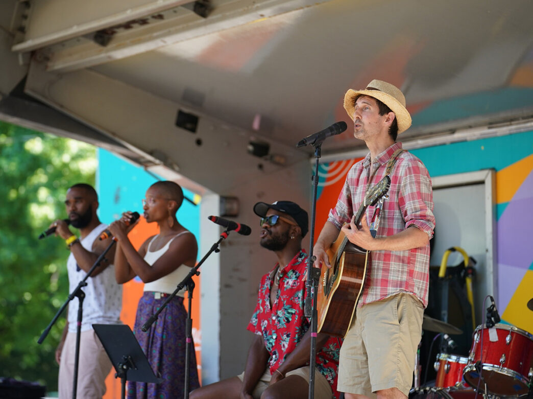 Musicians performing on an outdoor stage at the Kidchella Music Festival, featuring singers, a guitarist and a drummer against a colorful backdrop.