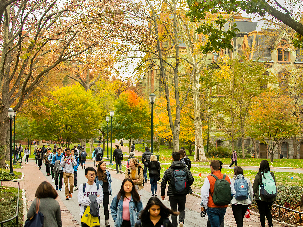 Students walk along a brick walkway through UPenn's campus during fall. Trees line the walkway and have yellow, orange and green leaves.