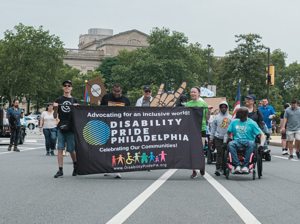 People holding a black banner lead the Disability Pride Philadelphia Parade down a street. People behind them hold cardboard signs in support.