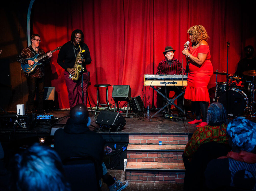 A pianist, guitarist and saxophonist play on stage while a person in a red dress sings into a microphone. A red curtain hangs behind the stage.