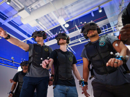 A group of players in motion-tracking suits, VR headsets and vests engage in a fully immersive virtual reality experience at Sandbox VR, surrounded by futuristic tech.