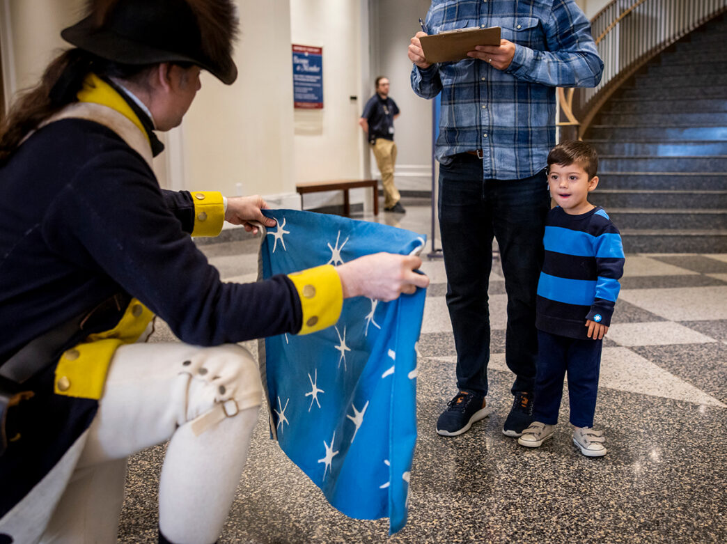 A Revolutionary War impersonator kneels down and holds up a replica of Washington's Headquarters Flag in front of a child visiting the Museum of the American Revolution in Philadelphia.