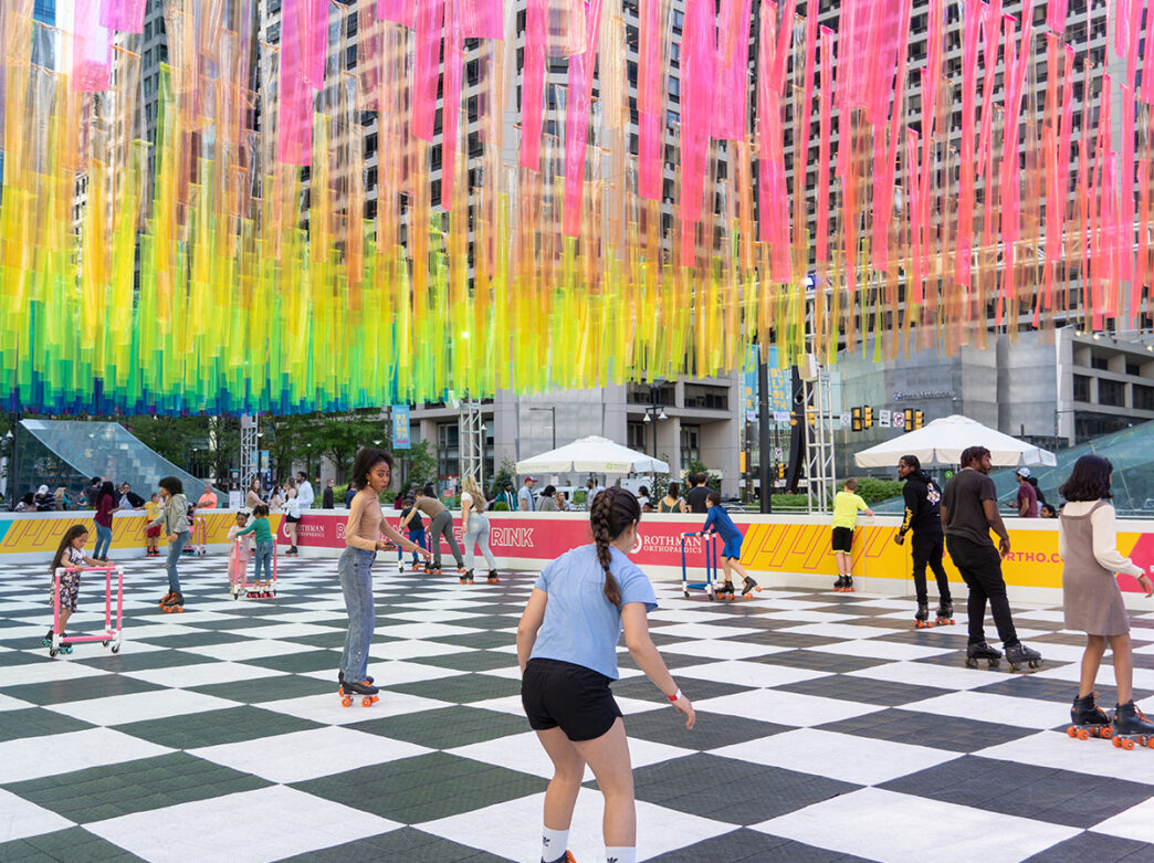 People rollerblade on a black and white roller rink at Philadelphia City Hall. Colorful streamers hang from above.