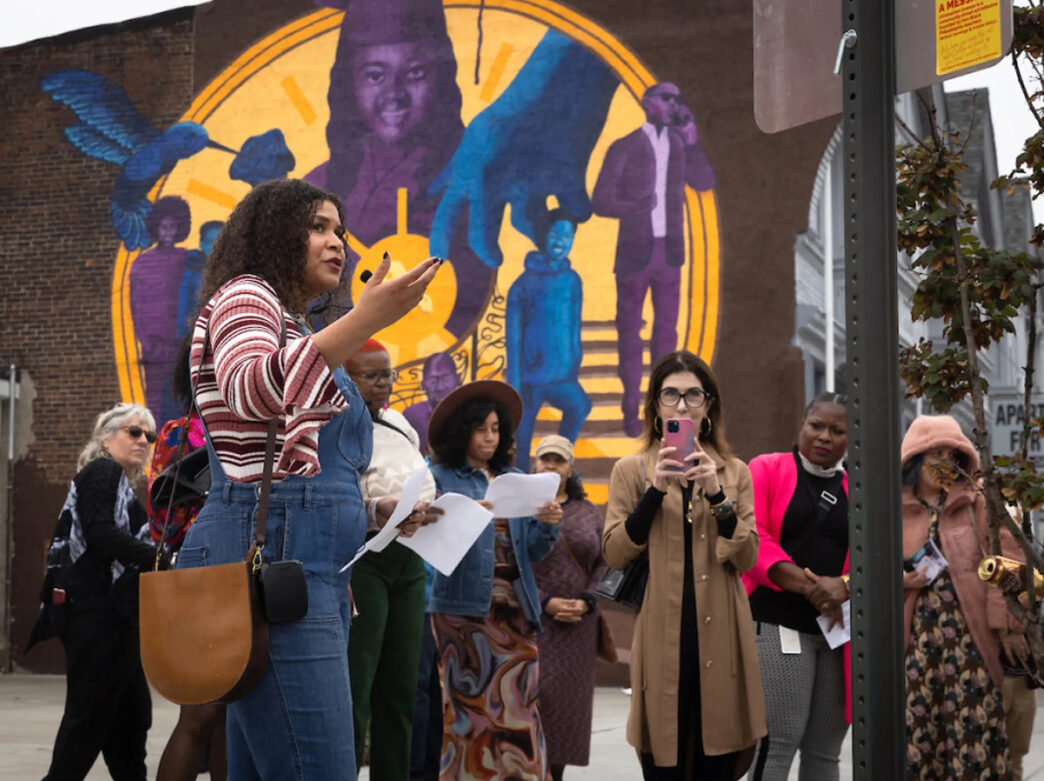 A tour guide leads a group on a walking tour. The group stops in front of a large mural on the side of the building while the guide talks.