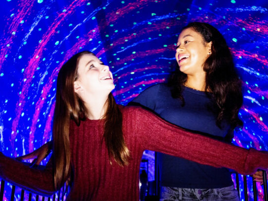 Dos amigos se ríen y se agarran a la barandilla mientras caminan por un vibrante y vertiginoso túnel de luces de neón e ilusiones ópticas en el Museo de las Ilusiones de Filadelfia.